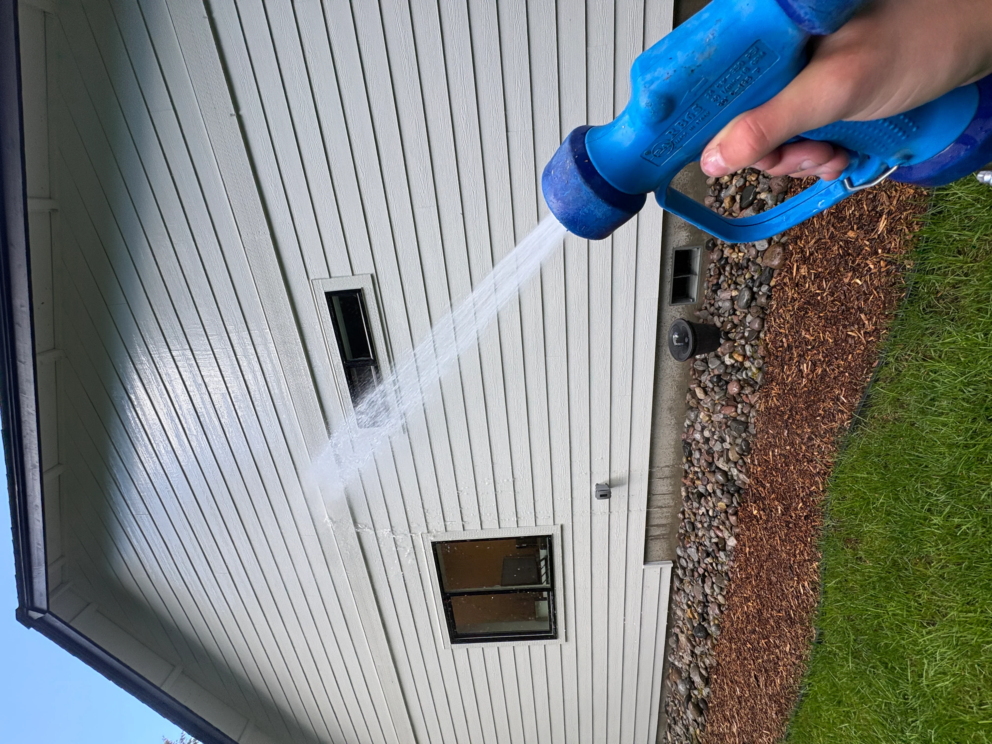 Pressure washing vinyl siding with blue nozzle, white ribbed panels and gravel foundation visible