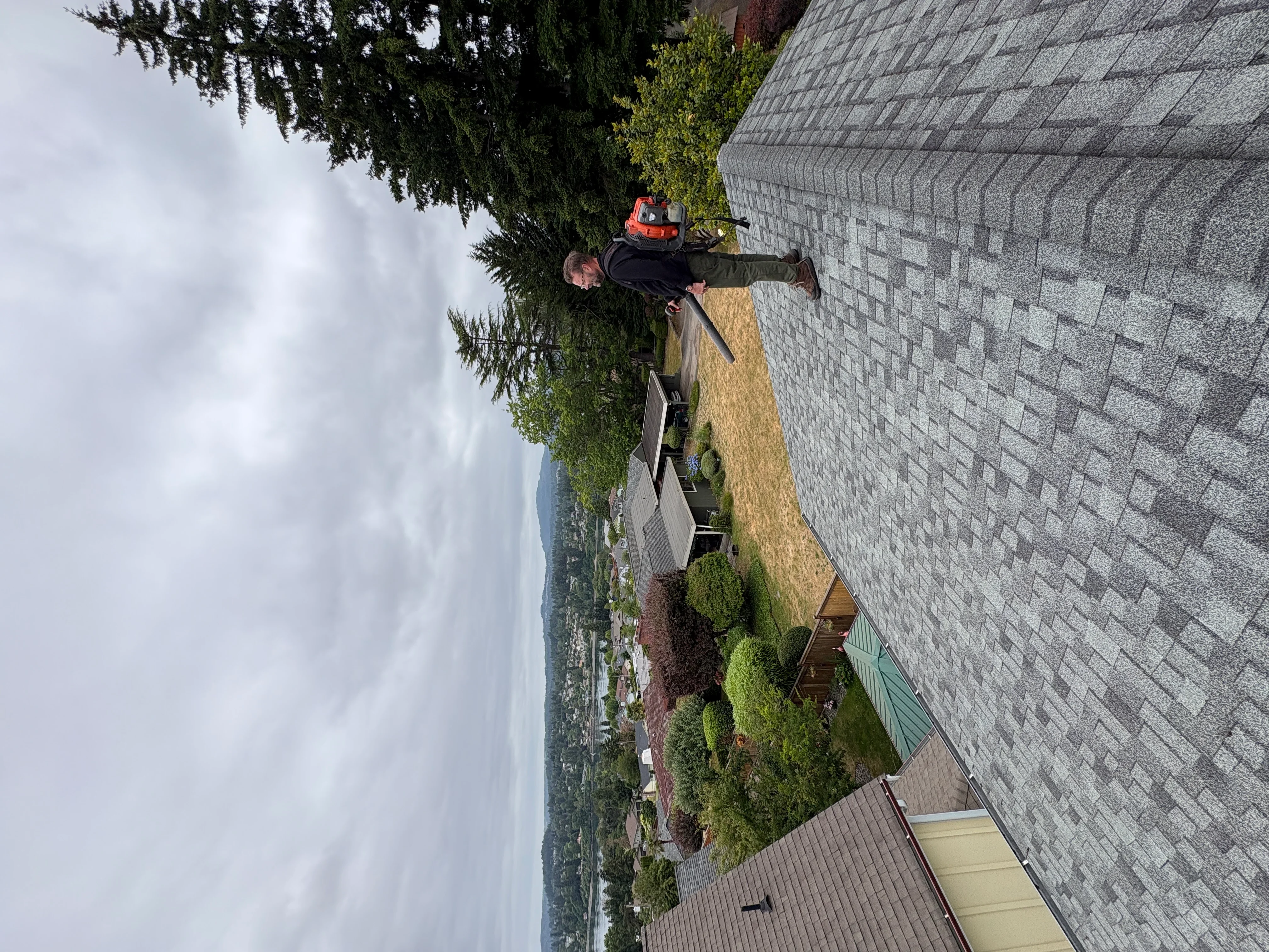 Technician cleaning gutters and roof debris from two-story home with gray shingle roof